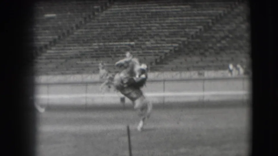 A rider on horseback continues across the Denver rodeo arena, partially framed by dark film edges and a foreground post.