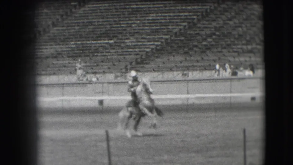 A horse and rider pass across the stadium field, with rows of empty seats and a few distant spectators behind.