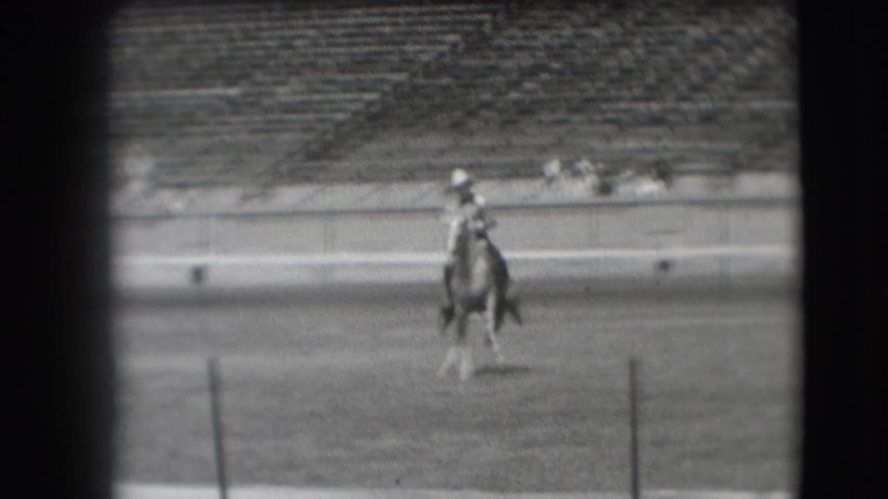 The mounted rider appears closer to the camera on the dirt field, framed by stadium seating in the background.