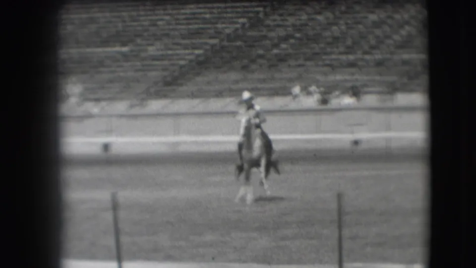 The mounted rider appears closer to the camera on the dirt field, framed by stadium seating in the background.