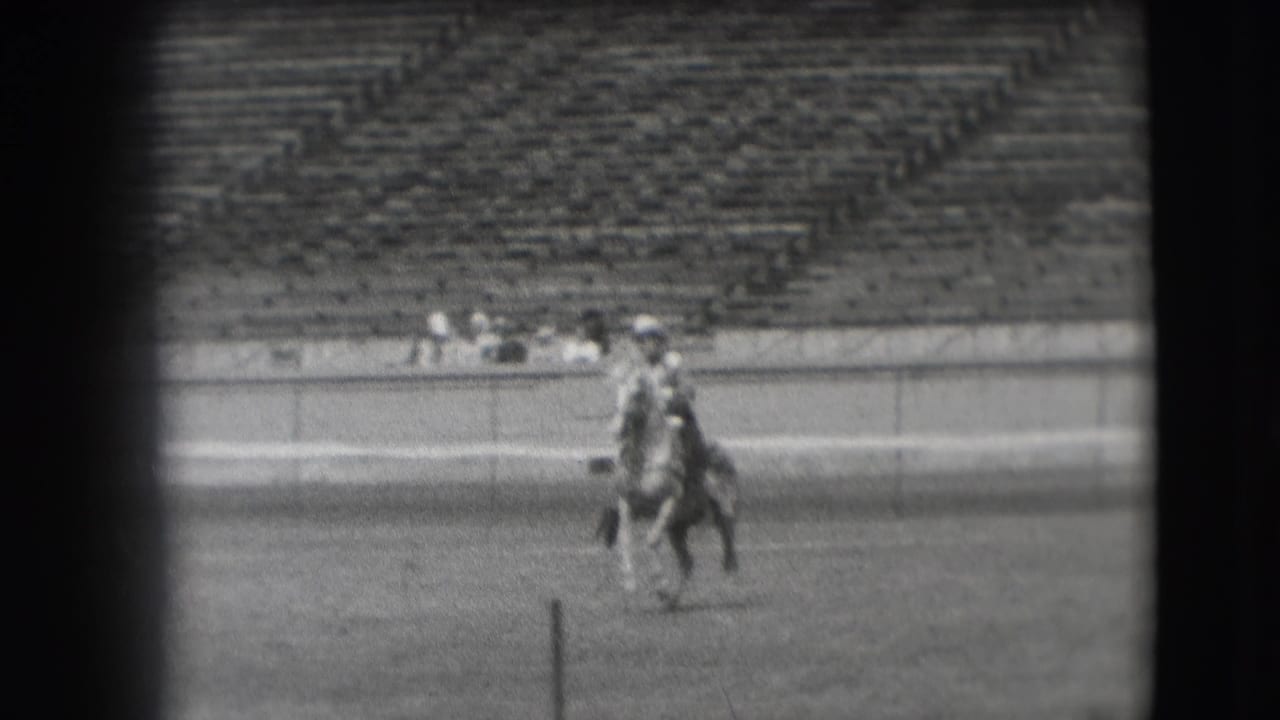 A rider on horseback continues across the field, partially aligned with a foreground post and distant stadium spectators.