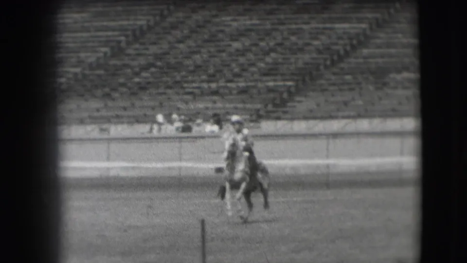 A rider on horseback continues across the field, partially aligned with a foreground post and distant stadium spectators.