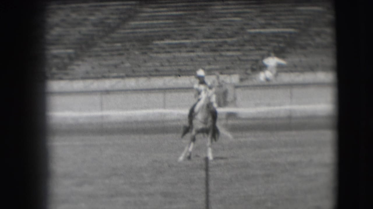 The rider continues across the Denver stadium arena on horseback, filmed from a distant spectator viewpoint in black and white.
