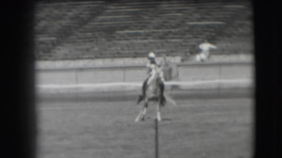 The rider continues across the Denver stadium arena on horseback, filmed from a distant spectator viewpoint in black and white.