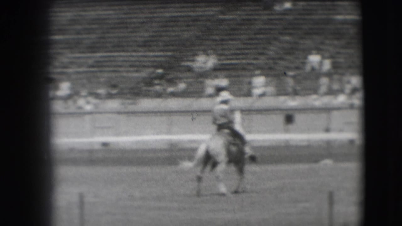 Mounted rider in a cowboy hat moves across a dirt arena with spectators in the background.