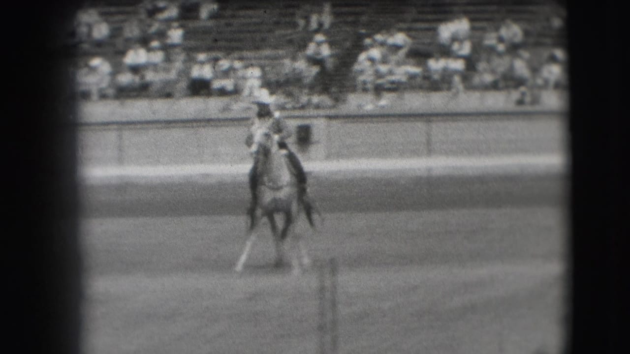 Cowboy on a light-colored horse in a dirt rodeo arena with spectators in bleachers.
