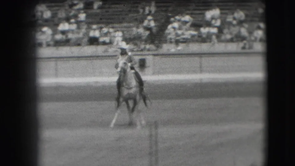 In 1941 Denver footage, a cowboy rides a light-colored horse across a dirt arena before a seated bleacher crowd.