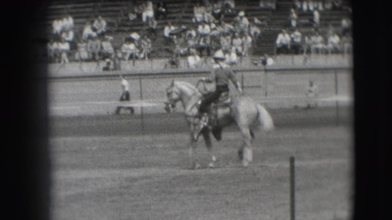 A cowboy in a dark outfit rides a light-colored horse across the Denver arena, filmed from a distant spectator viewpoint.