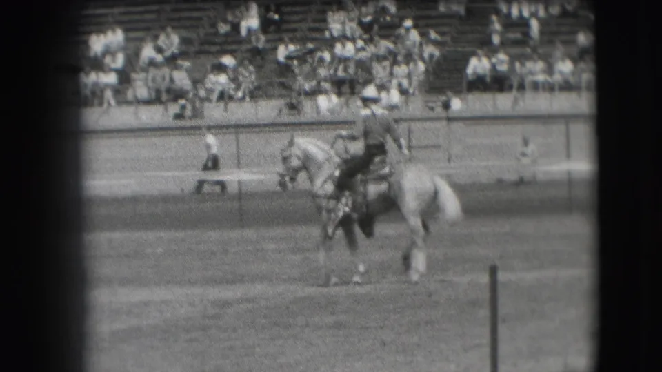 A cowboy in a dark outfit rides a light-colored horse across the Denver arena, filmed from a distant spectator viewpoint.