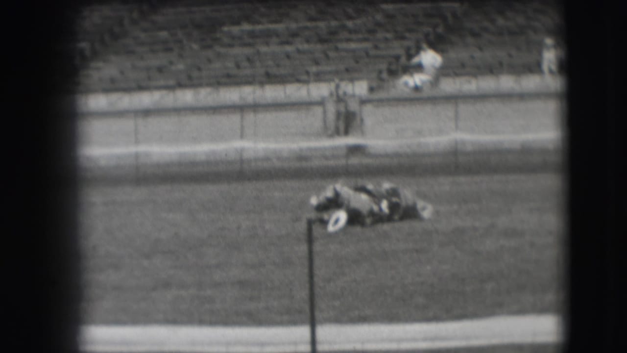 Horse lying on the grass beside a vertical pole, with empty stadium seating in the background.