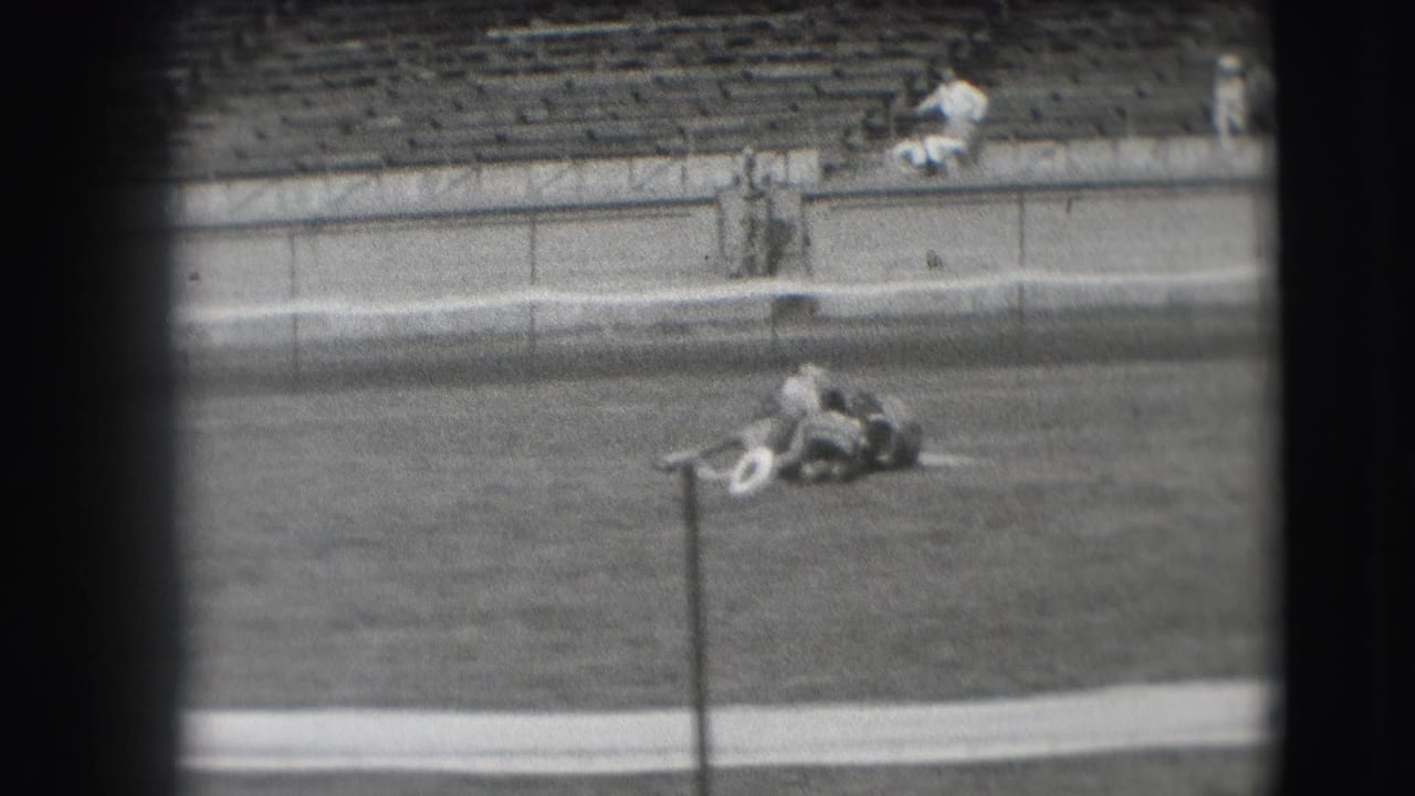 From a distant 8mm spectator viewpoint in Denver, the horse remains down on the arena grass near a pole with stadium seating beyond.