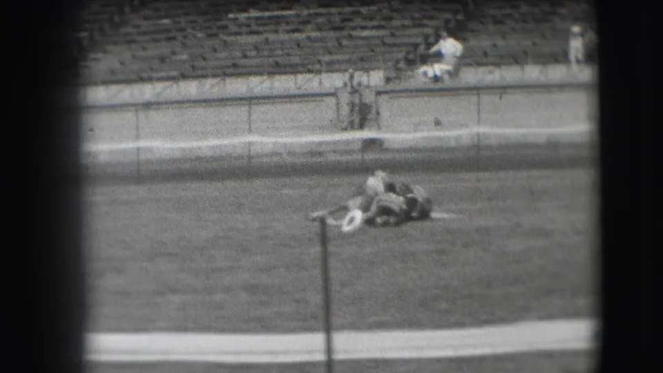 From a distant 8mm spectator viewpoint in Denver, the horse remains down on the arena grass near a pole with stadium seating beyond.