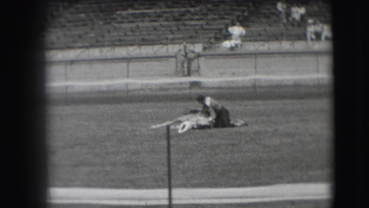 1941 Denver home-movie frame of a cowboy beside a lying horse in a grassy arena, seen with sparse spectators and bleachers behind.