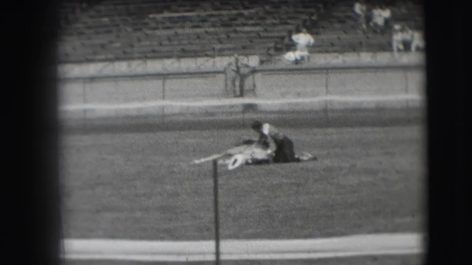 1941 Denver home-movie frame of a cowboy beside a lying horse in a grassy arena, seen with sparse spectators and bleachers behind.