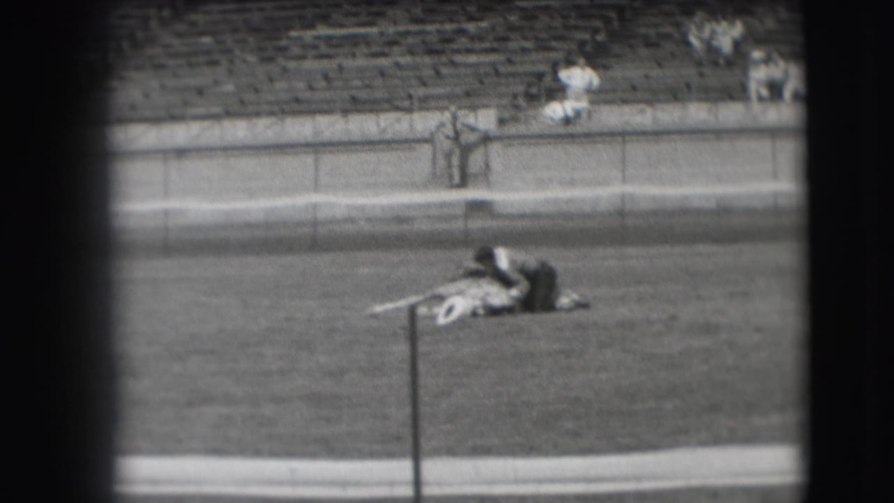 Man lies across a horse on a grass field with bleachers behind.