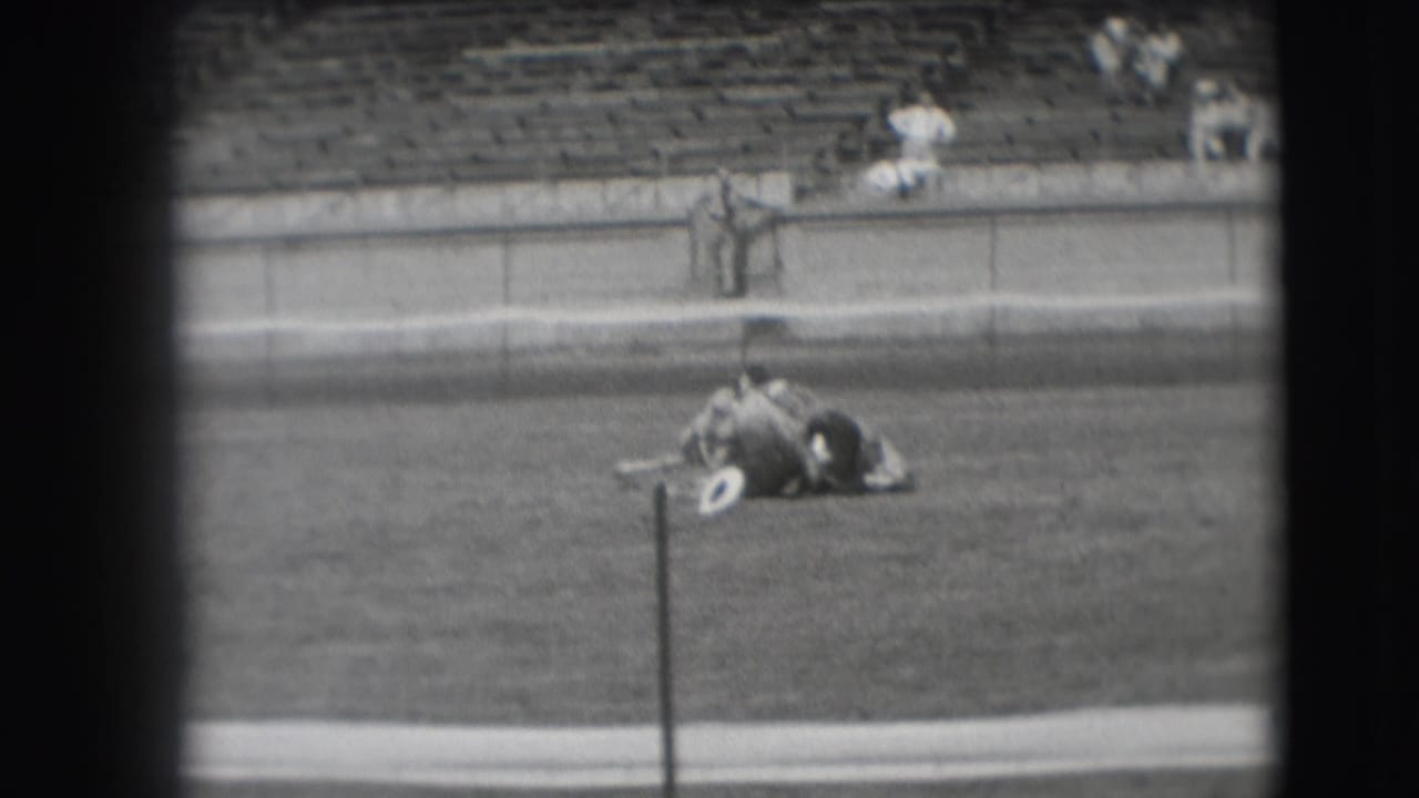 The horse lifts from the grass as the man stays across its back, with bleachers and a few spectators in the background.