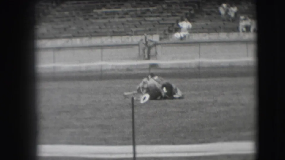 The horse lifts from the grass as the man stays across its back, with bleachers and a few spectators in the background.