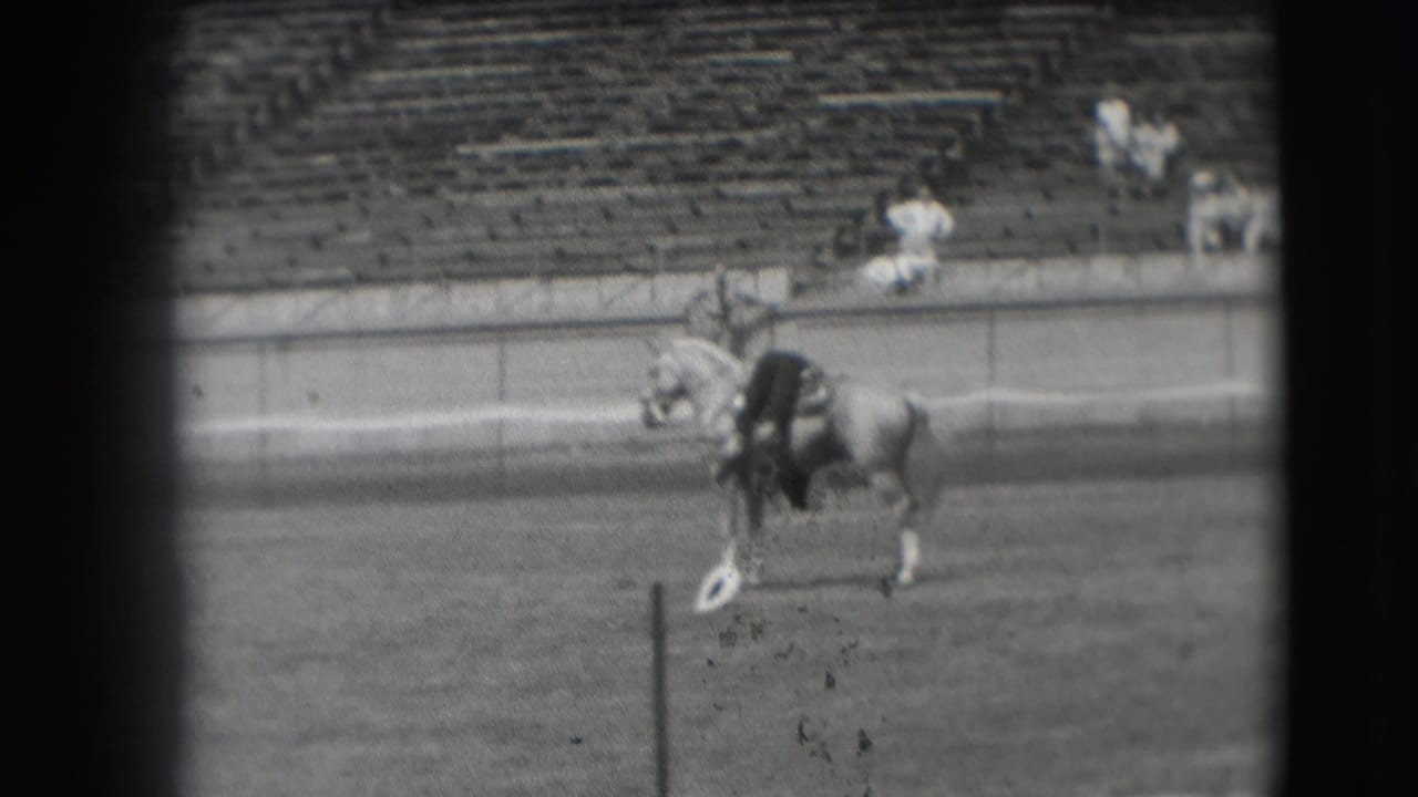 The horse and rider continue across the Denver arena, passing a small white marker while distant spectators sit in the stands.