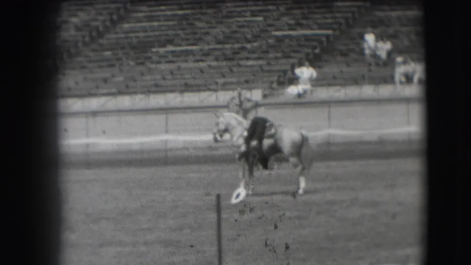 The horse and rider continue across the Denver arena, passing a small white marker while distant spectators sit in the stands.