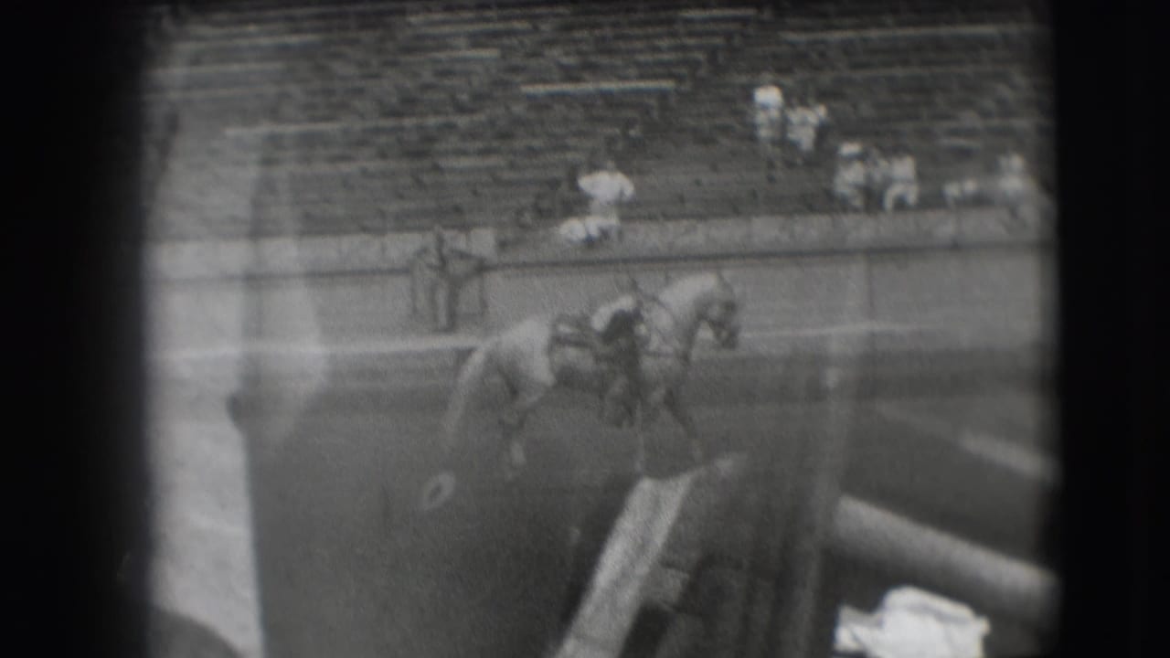 The mounted rider travels along the arena near the rail, with blurred foreground obstruction and mostly empty Denver bleachers behind.