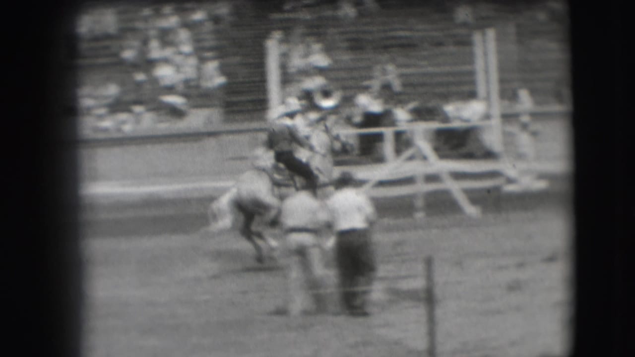 Cowboy rides a bucking horse in a rodeo arena with spectators in the bleachers.