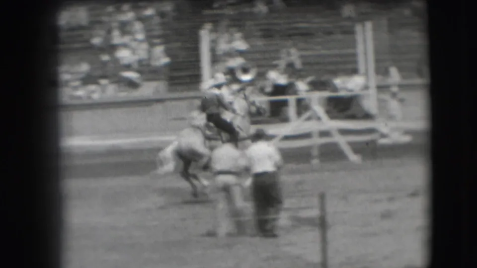 In 1941 Denver rodeo footage, a cowboy in a wide-brimmed hat rides a bucking horse while an arena attendant stands nearby.