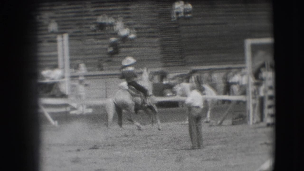 Mounted cowboy continues riding near the arena rail as an attendant watches.