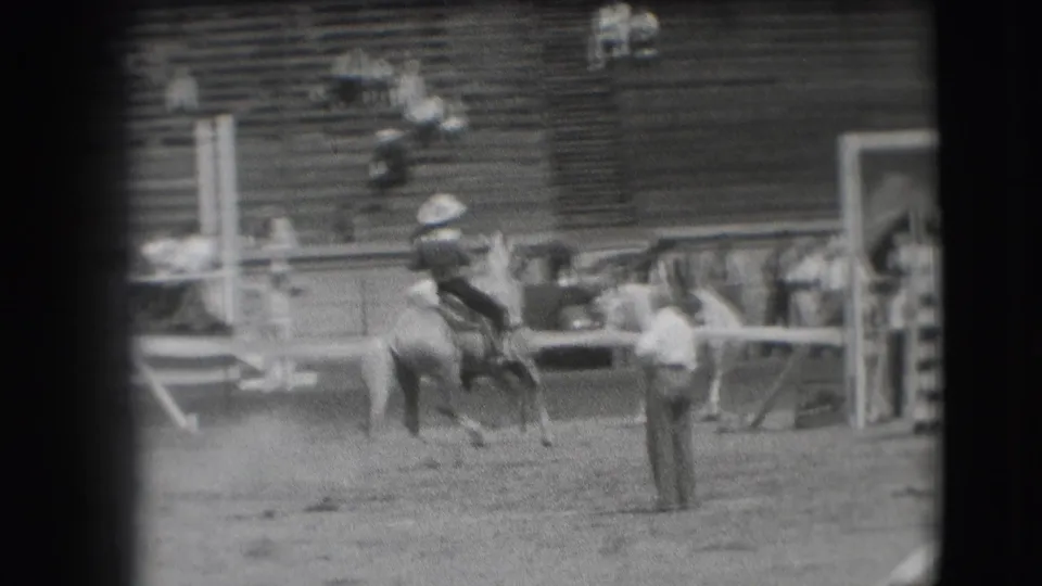 The cowboy and bucking horse move across the Denver rodeo arena, with a light-shirted attendant and bleachers visible behind them.