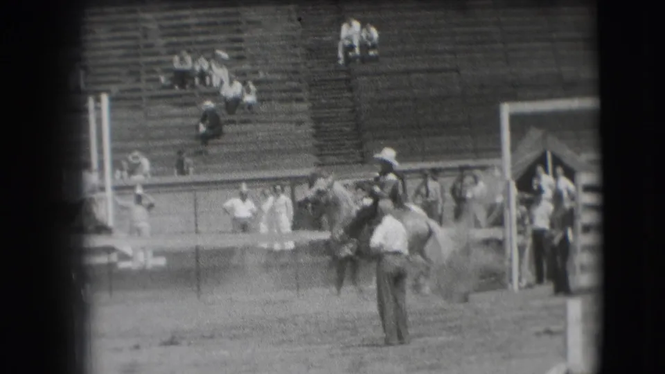 A rodeo rider on horseback appears near the gate area, with arena staff, rail fencing, and bleacher spectators in the background.