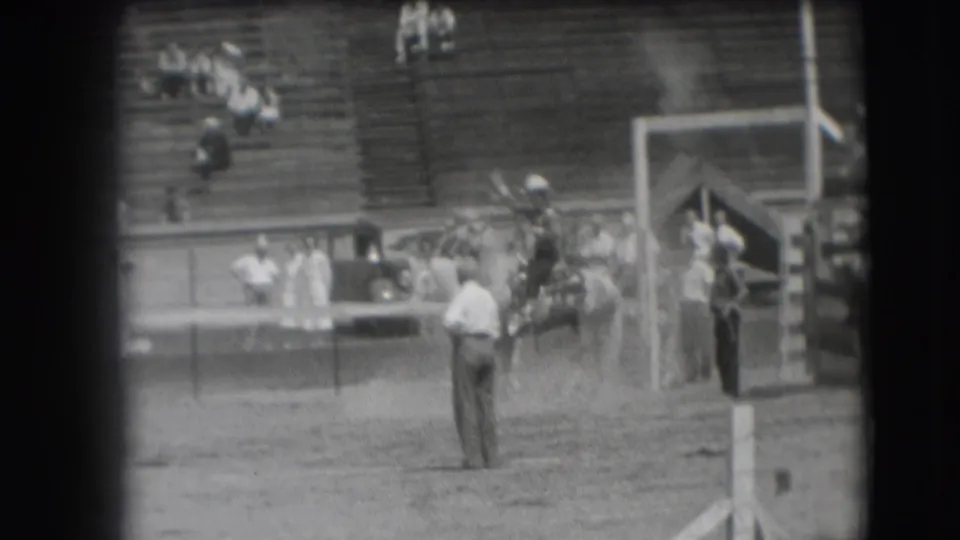 The brief rodeo sequence ends with the horse and rider near the fence, an attendant in the foreground, and spectators seated in the bleachers.