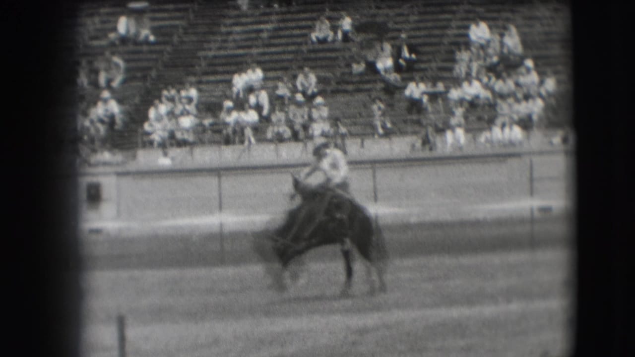 Cowboy riding a bucking horse in an arena with spectators in bleachers behind.