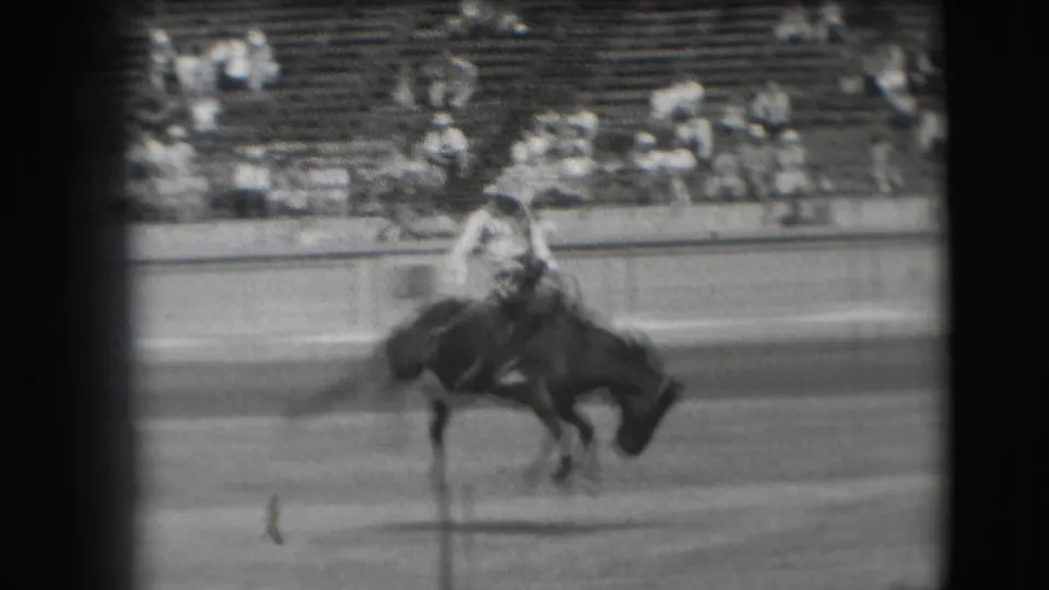 A cowboy leans over a bucking horse in the Denver rodeo arena, captured in grainy black-and-white 8mm footage.