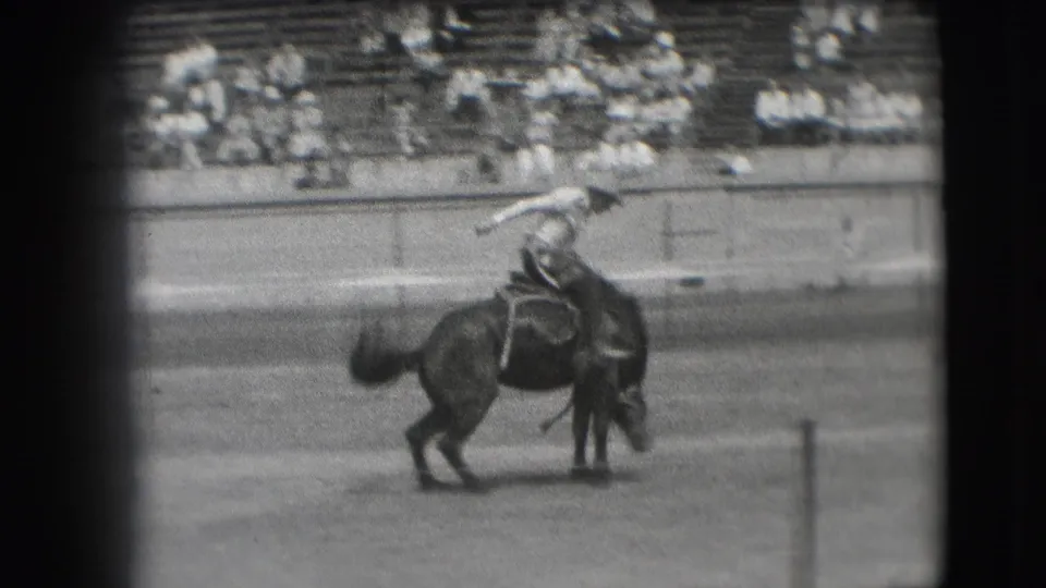 The rider extends an arm as the bucking horse twists in the 1941 Denver rodeo arena, with spectators visible behind.