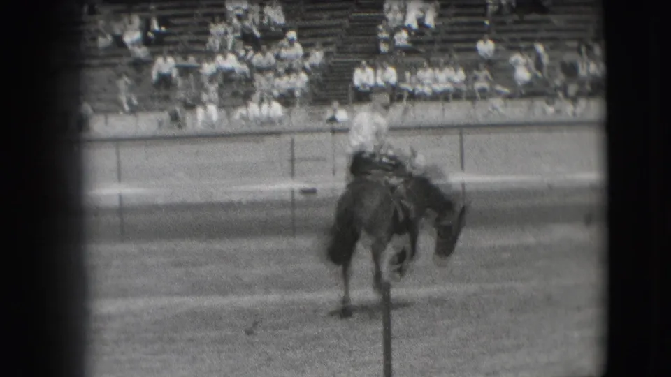 1941 Denver rodeo frame showing a cowboy on a dark horse in the dirt arena, with blurred bleachers and spectators in the background.