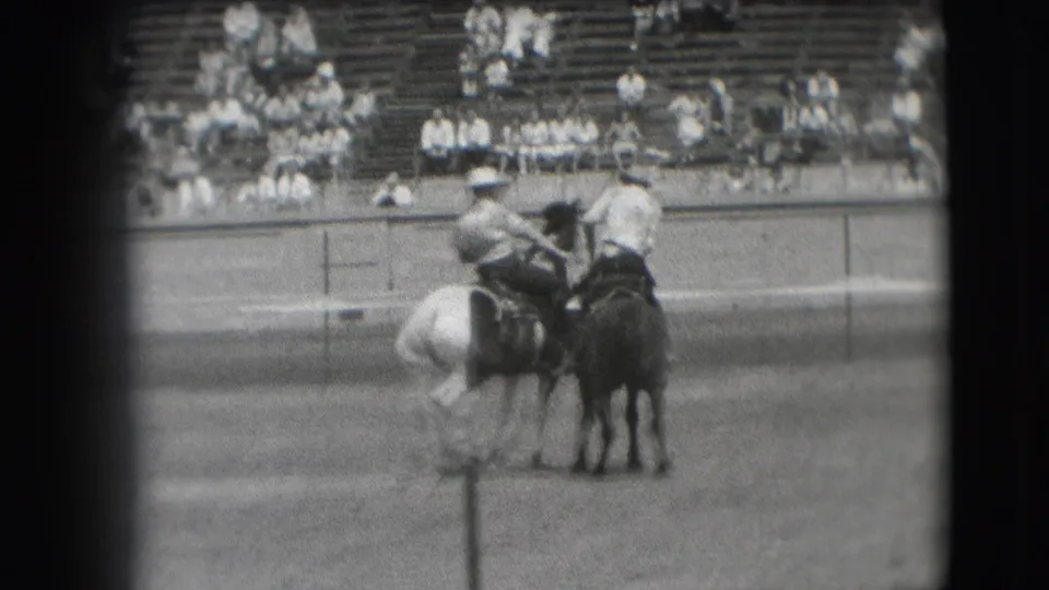 Two cowboys on horseback appear near each other in the Denver rodeo arena, with bleachers and spectators behind them.