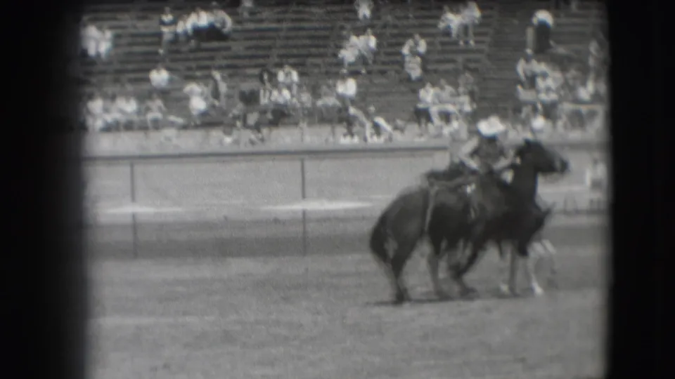 A mounted cowboy on a dark horse moves across the Denver rodeo arena, with spectators seated in bleachers beyond the rail.