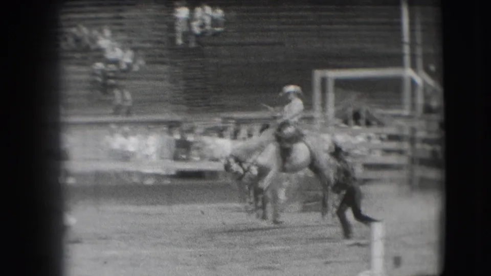 A cowboy rides a bucking horse in the Denver rodeo arena, with wooden chutes and bleachers visible in the background.