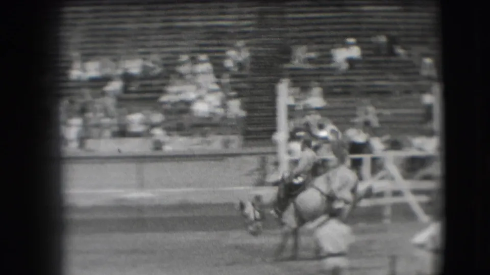 The rider and horse move across the Denver rodeo arena near the rail, seen through soft black-and-white 8mm film grain.
