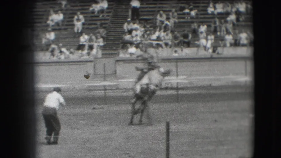 A cowboy rides a bucking horse in the Denver arena as a man on foot stands nearby and spectators watch from the bleachers.