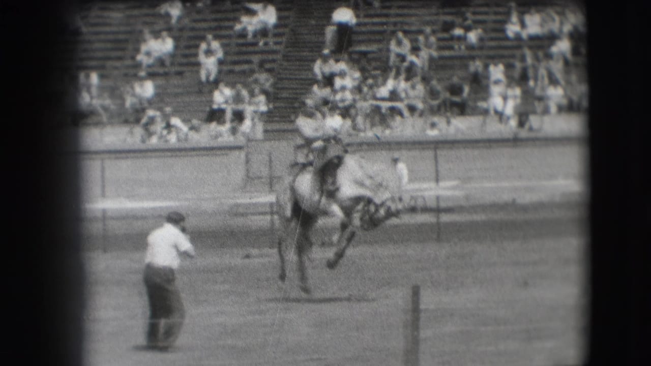 Cowboy rides a bucking horse in a rodeo arena as a man watches from the dirt.