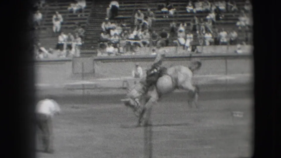 The rodeo horse drops and twists as the cowboy struggles to stay seated, with bleachers and a nearby observer in view.