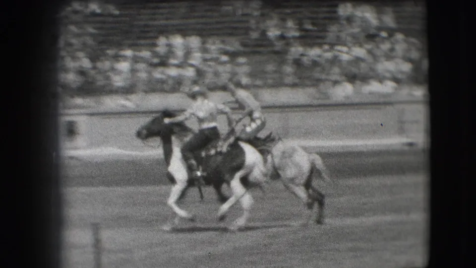 In the stated 1941 Denver rodeo setting, a cowboy leans low during a horseback trick as spectators sit in the grandstand.