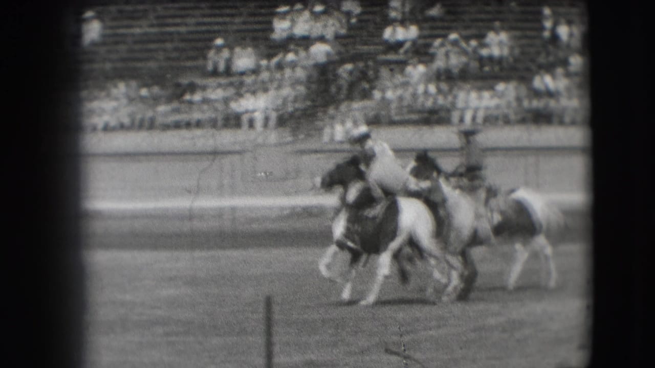 Two mounted riders pass through the arena during the Denver rodeo sequence, with spectators visible behind the rail.