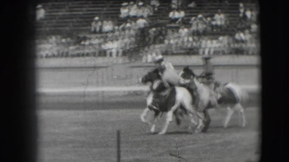 Two mounted riders pass through the arena during the Denver rodeo sequence, with spectators visible behind the rail.