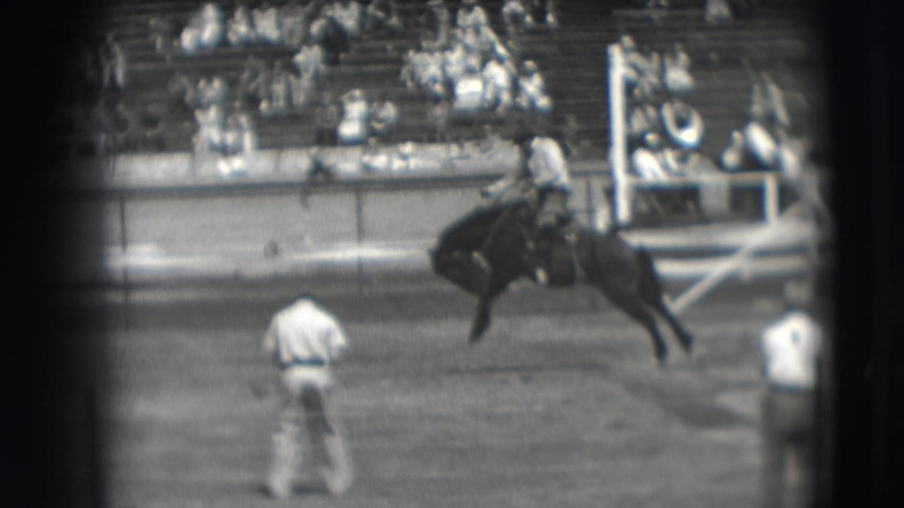 Horse bucks across the arena while a rider holds on and men stand nearby.