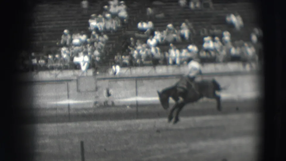 The rider appears off balance as the bucking horse kicks near the rodeo barrier in the supplied 1941 Denver home-movie frame.
