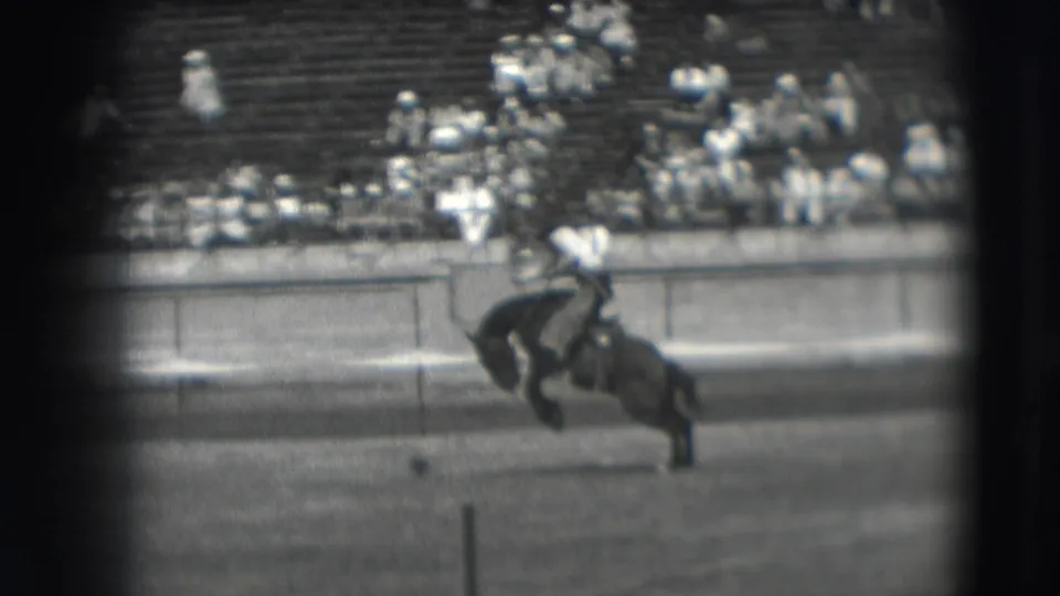A rider continues holding on during the Denver rodeo sequence, with the bucking horse framed against bleachers and arena fencing.