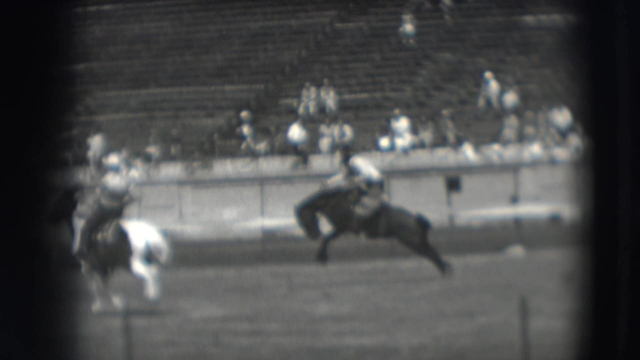 Mounted riders gallop across a stadium field with spectators in bleachers behind them.