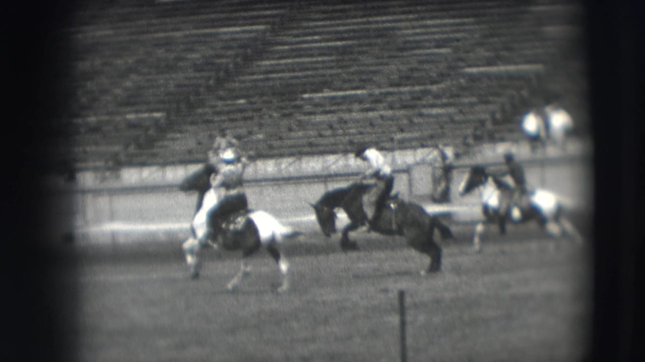 Two riders on horseback move through a rodeo arena with stadium seating behind them.
