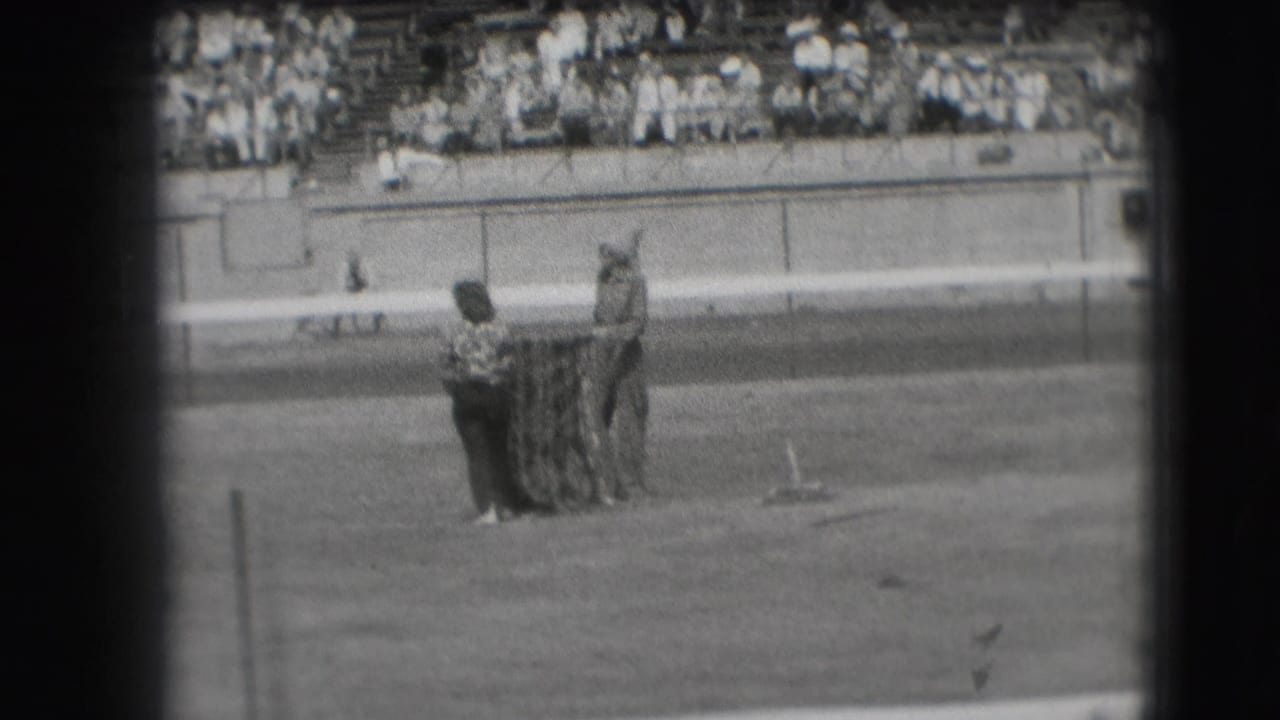 Two horseshoes players stand near a pit on a field with spectators in bleachers behind them.